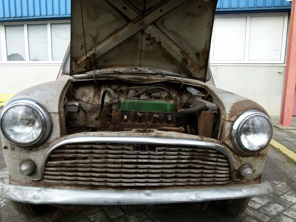 Voiture Mini rouillée avec capot ouvert révélant moteur, devant un bâtiment industriel.